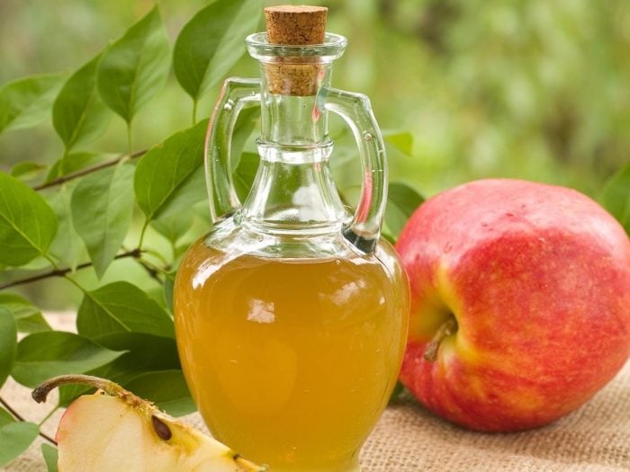 A jar of apple cider vinegar against a green background with whole and halved fresh apples and leaves