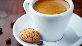 Close-up of an espresso shot in a cup with saucer containing a cookie on a wood surface scattered with coffee beans.