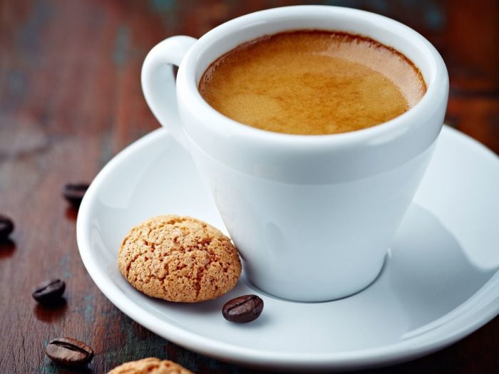 Close-up of an espresso shot in a cup with saucer containing a cookie on a wood surface scattered with coffee beans.