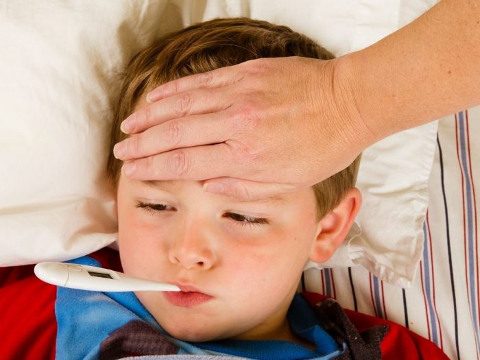A kid suffering from fever lying on the bed with a thermometer in his mouth & a family member checking the body temperature