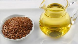 A white bowl filled with flaxseed and a jar filled with flaxseed oil kept on a white wooden table