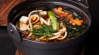 A soup pot of japanese miso soup of tofu, green onions, carrots, and udon noodles on a wooden mat
