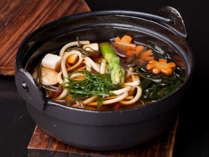 A soup pot of japanese miso soup of tofu, green onions, carrots, and udon noodles on a wooden mat