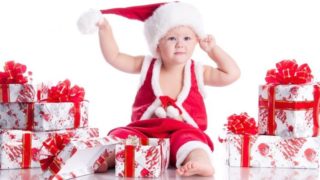 A baby dressed up in red clothes and Christmas cap sitting in between gifts against a white background