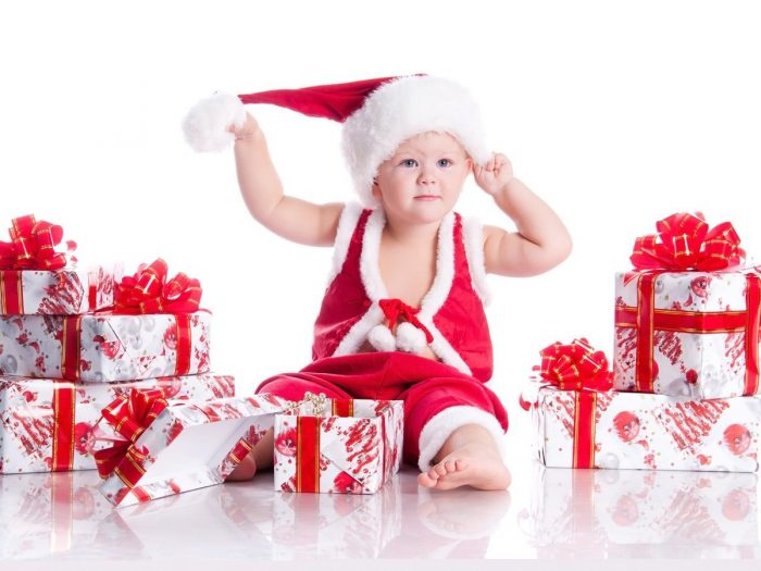 A baby dressed up in red clothes and Christmas cap sitting in between gifts against a white background