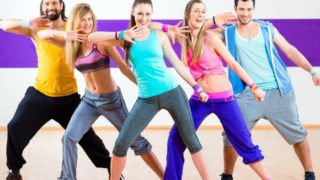 A group of smiling young people doing zumba on a wooden floor in a fitness center
