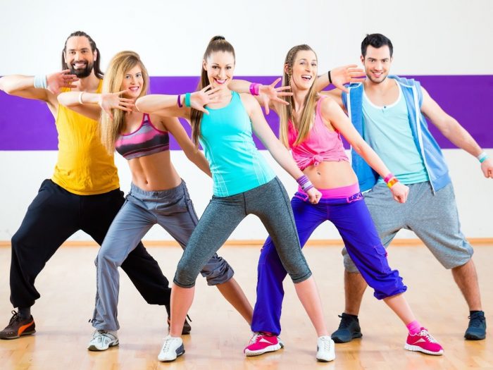 A group of smiling young people doing zumba on a wooden floor in a fitness center