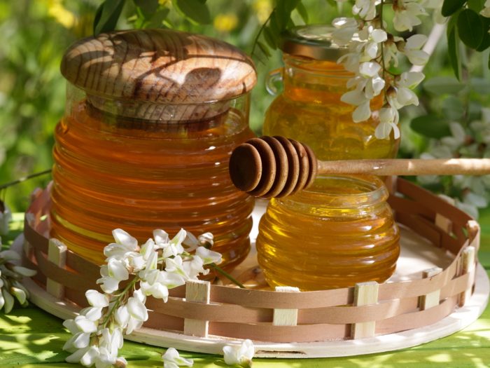 A jar and of acacia honey with a wooden stirrer and white flowers on a wooden tray