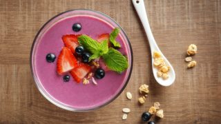 Flat-lay shot of a purple smoothie with strawberries and black grapes on a wooden surface next to a spoon and some nuts