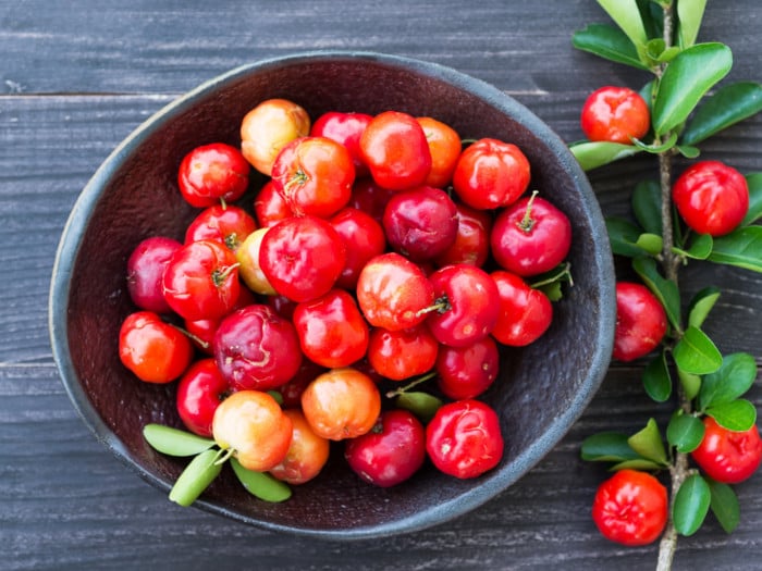 a bowl of fresh red acerola cerejas and leaves on a wooden table