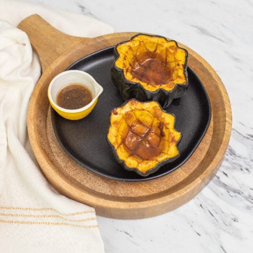 Roasted acorn squash and maple syrup on a black plate on a wooden tray, placed on a white countertop