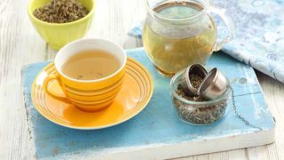 A cup of agrimony tea kept near a bowl of agrimony dried leaves and a pitcher of the tea, on a blue platform