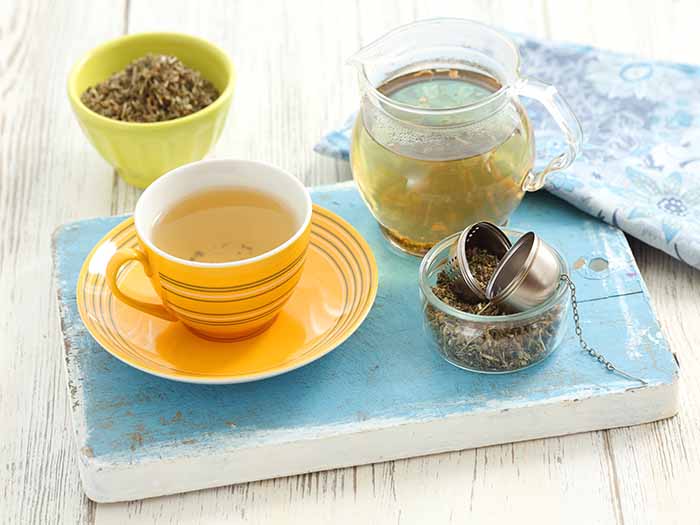 A cup of agrimony tea kept near a bowl of agrimony dried leaves and a pitcher of the tea, on a blue platform