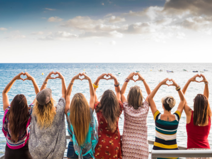 Seven woman facing a beach with their hands above their head making a heart individually