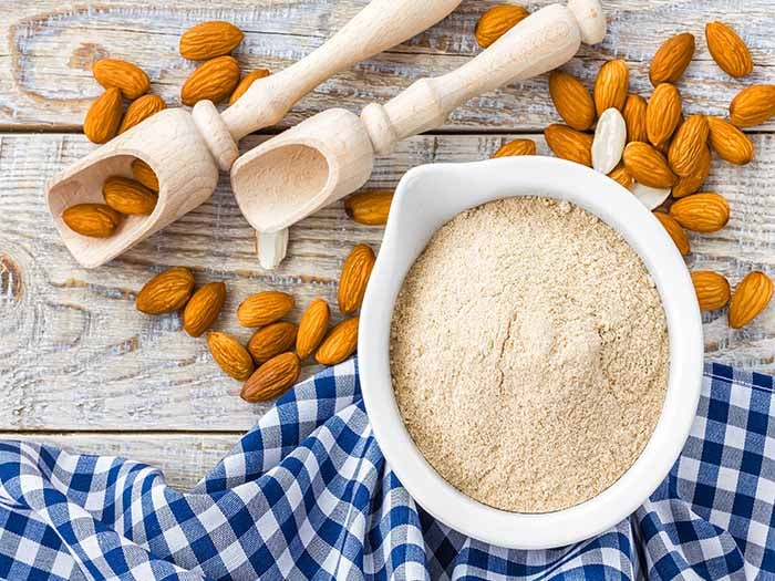 A flat lay picture of a bowl of almond flour kept next to almonds on a wooden table