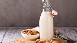 A white bowl of shelled almonds, unshelled almonds, almond milk bottle, and spoons on a grey background