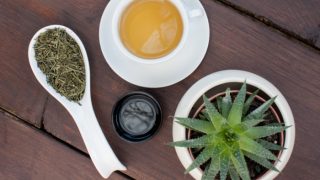 A flat lay picture of a cup of aloe vera tea kept beside a spoon of aloe vera dried leaves and a small cup of aloe vera gel