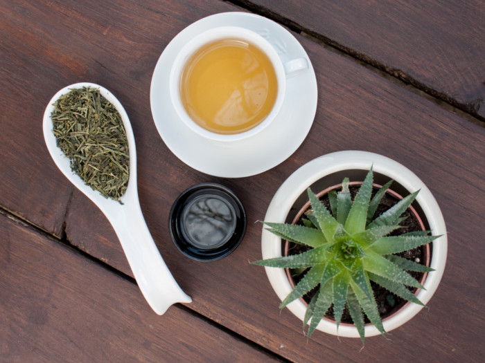A flat lay picture of a cup of aloe vera tea kept beside a spoon of aloe vera dried leaves and a small cup of aloe vera gel