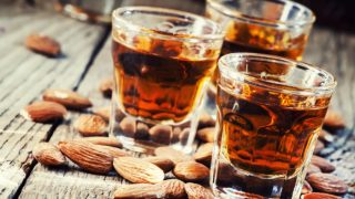 A close-up shot of three glasses of amaretto surrounded by almonds as it is placed on a wooden table