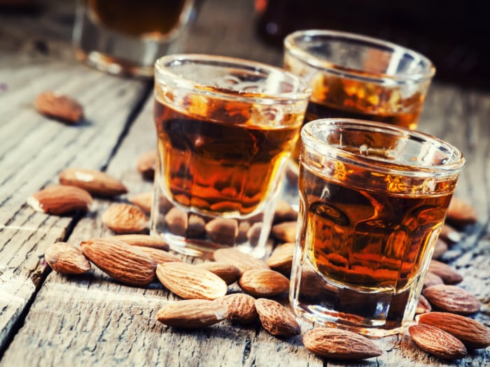 A close-up shot of three glasses of amaretto surrounded by almonds as it is placed on a wooden table