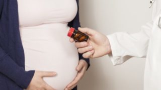 A hand holding a bottle of pills in front of a pregnant woman's stomach