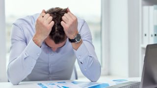 A man sitting at his work desk, looking down, with his hands pulling his hair
