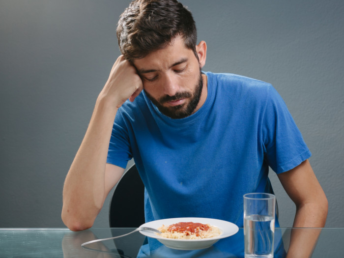A despondent looking man staring down at a plate of food placed alongside a glass of water in front of him