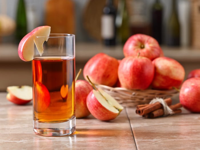 A glass of apple juice with sliced and whole apples as well as cinnamon sticks in the background