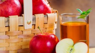 A glass of apple juice kept next to apples and a basket full of them, atop a table