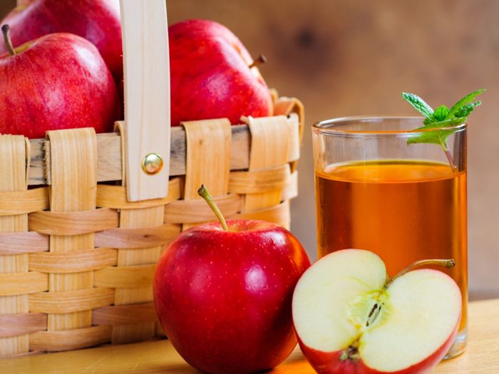 A glass of apple juice kept next to apples and a basket full of them, atop a table