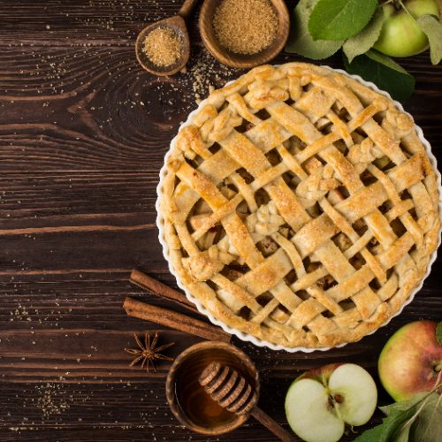 A flat lay picture of an apple pie and ingredients on a wooden background