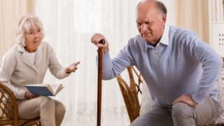 An older man trying to get out of his chair while an older woman looks at him with a worrisome look on her face