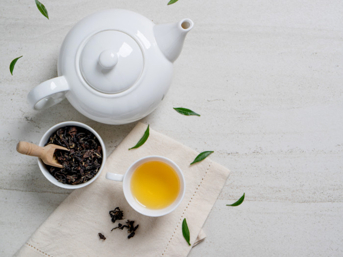 A flat lay picture of a cup of assam tea kept next to a kettle and assam tea leaves, on a white surface