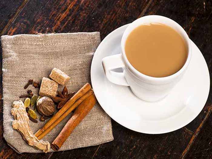 A cup of milky Assam tea, kept atop a wooden table, next to spices kept on a grey colored napkin