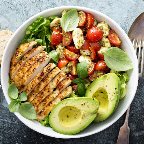 A white bowl of grilled chicken and avocado salad kept next to spoons and folks, atop a grey table
