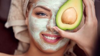 A young woman with an avocado face mask on her face smiling and holding half avocado