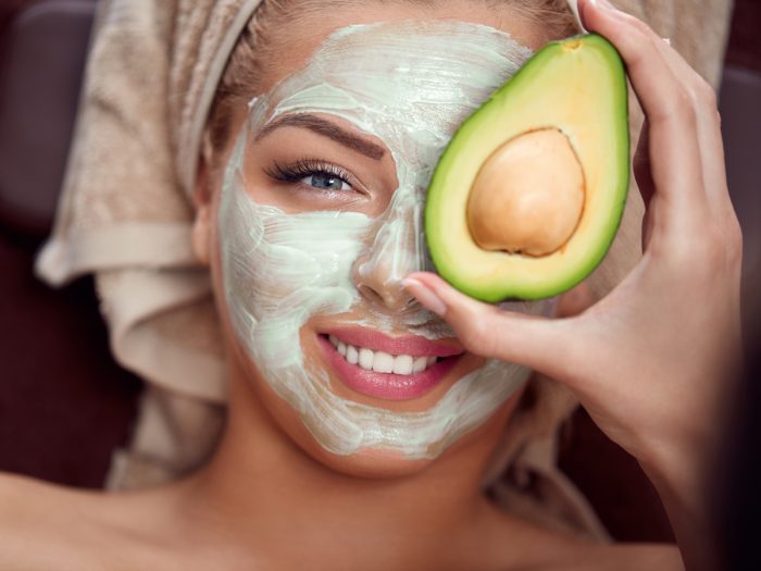 A young woman with an avocado face mask on her face smiling and holding half avocado