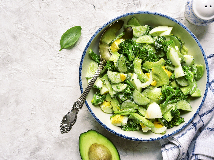 A flat lay picture of avocado salad with broccoli,cucumber and boiled eggs in a white vintage bowl over light slate,stone or concrete background