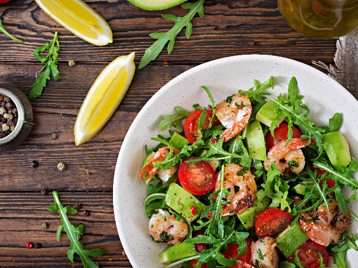 A close-up shot of a salad bowl with shrimp, tomato, avocado and arugula against a wooden background.