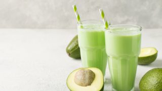 Two glasses of avocado smoothies placed atop a table next to avocados