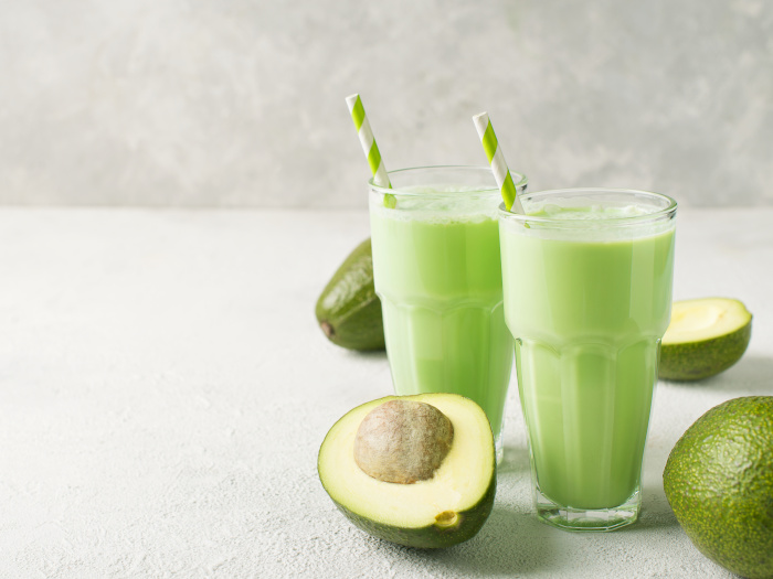 Two glasses of avocado smoothies placed atop a table next to avocados