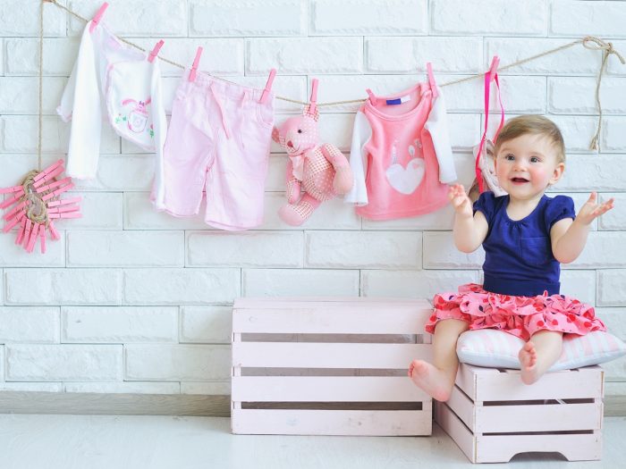 A baby sitting on a white wooden box and behind her on a white wall there are small baby clothes hanging