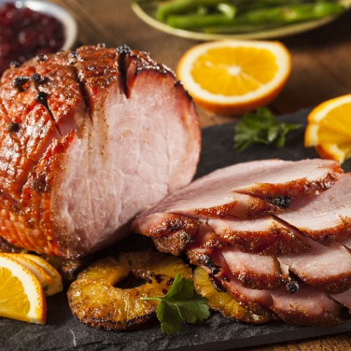 Roasted sliced Christmas ham on plate with fork, knife and festive decoration against a dark rustic background