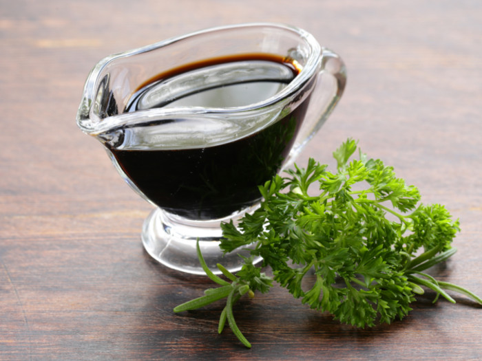 A sauce boat of a dark-colored liquid with a sprig of parsley on a wooden surface.