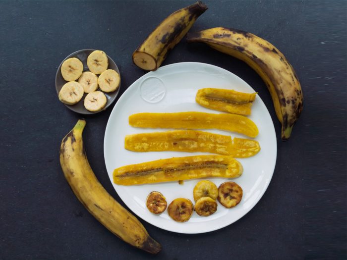 Flatline picture of a plate of plantain chips along with bowl banana slices and bananas on a slate background.