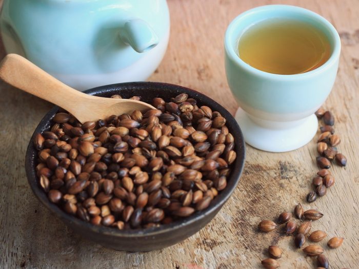 A ceramic bowl of roasted barley seeds with a teapot and cup of barley tea on a wooden table