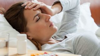 A woman looking ill lying on a bed, with pills and a glass of water on the side table