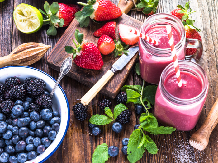 A flat-lay pic of berry smoothies kept atop a wooden platform along with a bowl of berries