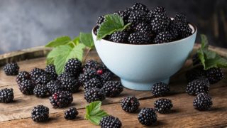 A white bowl of fresh blackberries with leaves on a wooden table