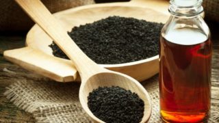 A wooden bowl and spoon of black cumin seeds with a bottle of black cumin oil on a jute cloth